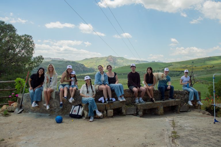 Students sit together on a stone wall overlooking rolling green hills under a blue sky.