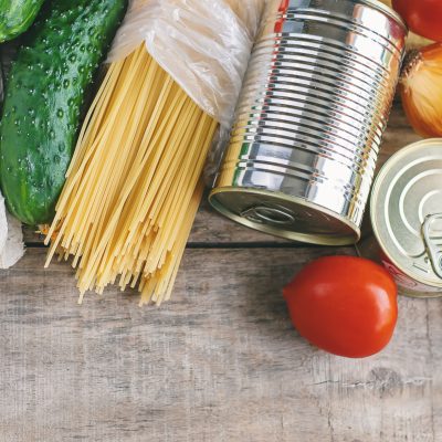 Overhead view of grocery staples including eggs, cucumbers, spaghetti, canned goods, onions, tomatoes, lemons, and cooking oil arranged on a wooden surface