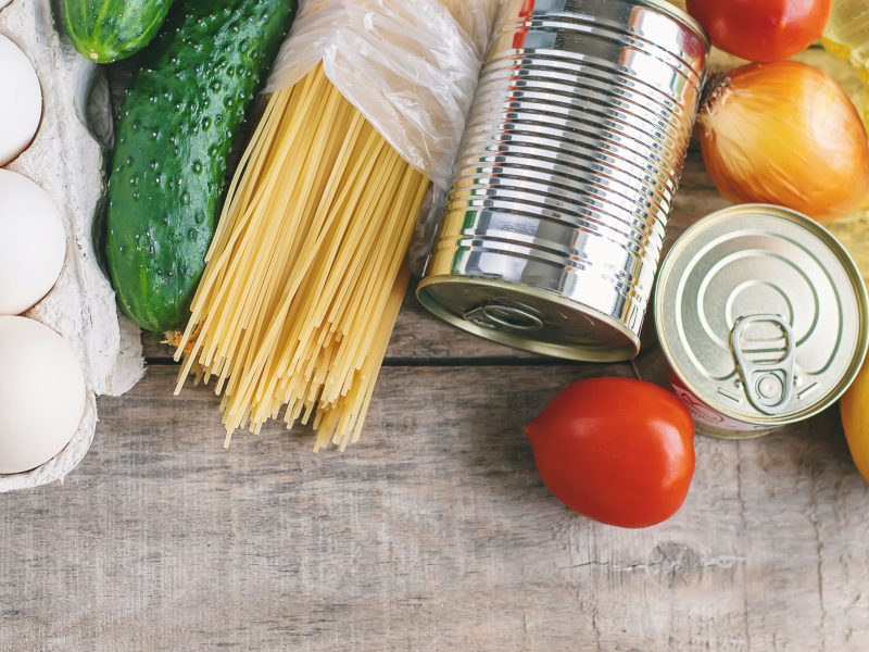 Overhead view of grocery staples including eggs, cucumbers, spaghetti, canned goods, onions, tomatoes, lemons, and cooking oil arranged on a wooden surface