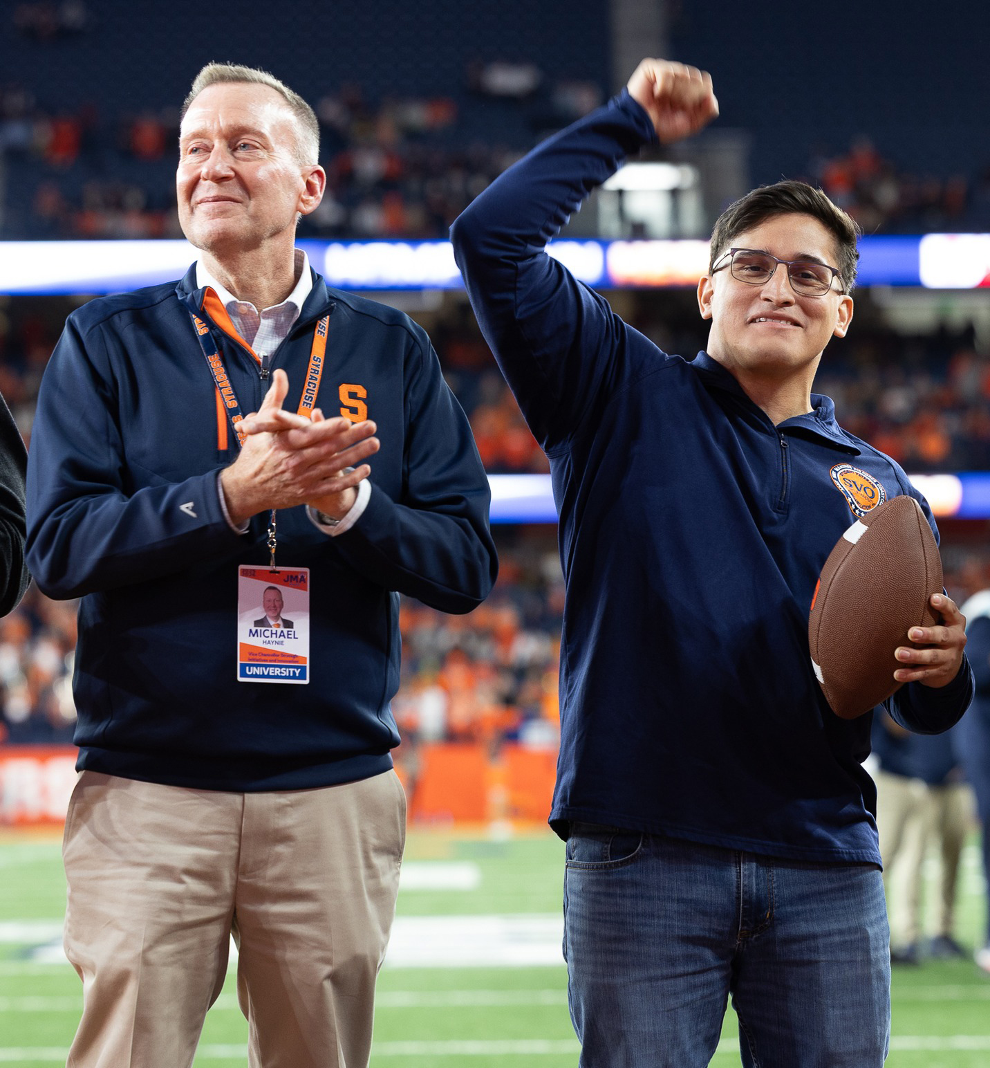 Person applauds beside a student holding a football and raising his fist in celebration at a stadium event.