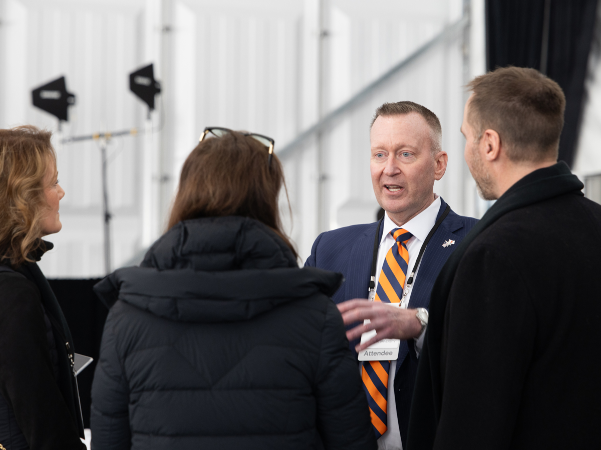 Person in an orange-striped tie speaks with a group of attendees at an outdoor event.