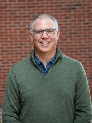 A person smiles while posing for a headshot in front of a red brick wall.
