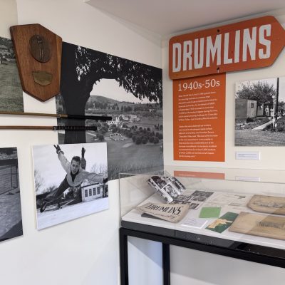 Museum exhibit about Drumlins in the 1940s-50s, featuring historic photos, golf clubs, and archival documents in a display case.
