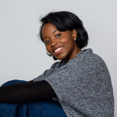 Person seated with knees drawn up, wearing jeans and a gray knit sweater, looking toward the camera against a plain light background.
