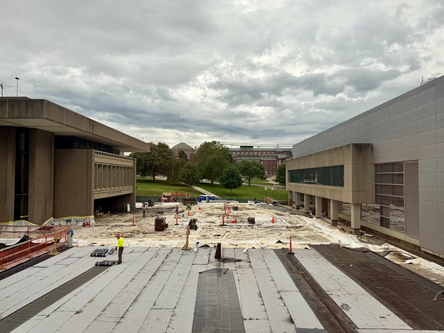 Construction work underway in the plaza between two campus buildings, with covered ground, construction materials, and workers under cloudy skies.