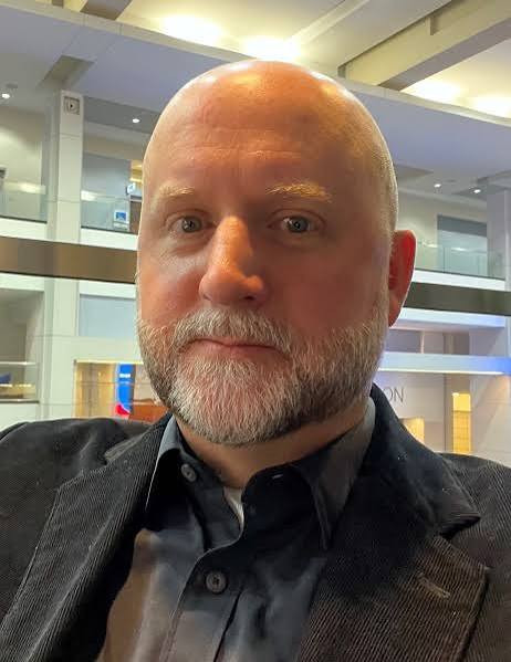 Head-and-shoulders portrait of a person in a dark blazer and button-down shirt in an indoor atrium.