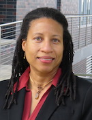 Professional portrait of a person in a dark blazer standing near a modern building with metal railings.