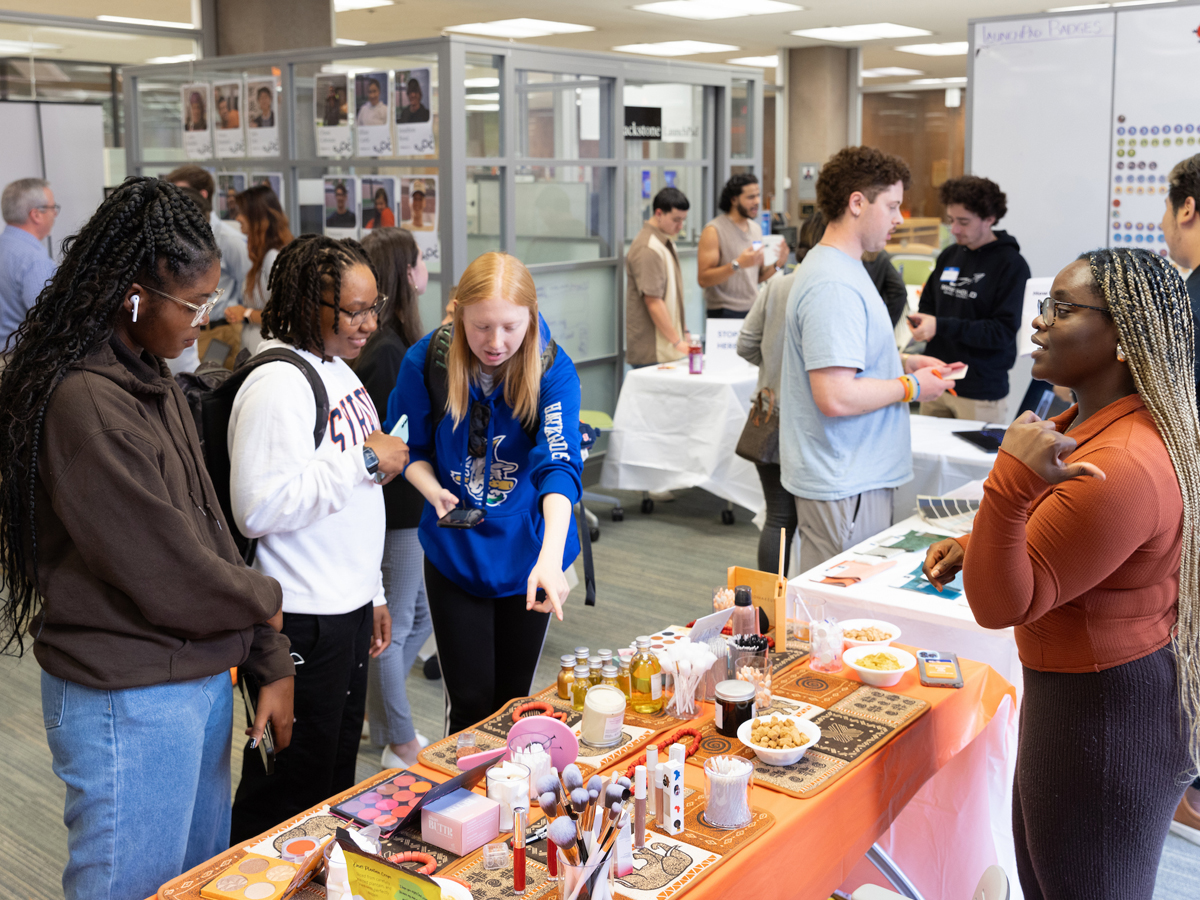 Students gather around a vendor table displaying beauty and skincare products at an indoor campus marketplace event, with a student entrepreneur explaining her offerings to three visitors.