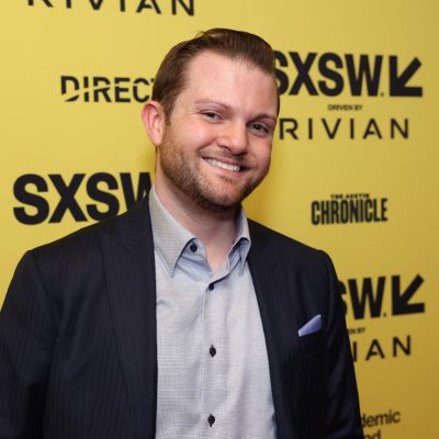 A person smiles in a sport coat and dress shirt while standing in front of a yellow SXSW promotional banner.