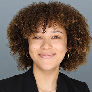 A professional headshot of a smiling woman with curly brown hair, wearing gold hoop earrings and a black blazer, photographed against a solid slate-blue background.