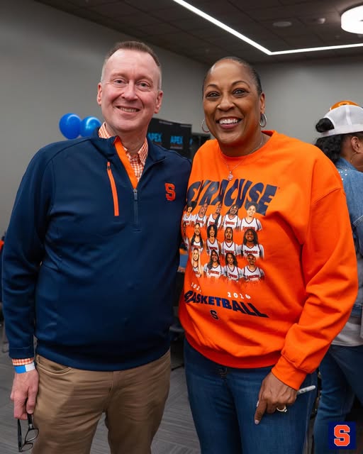 Two people smile for a photo indoors, with the woman wearing an orange Syracuse Basketball sweatshirt featuring team photos, and blue balloons visible in the background.