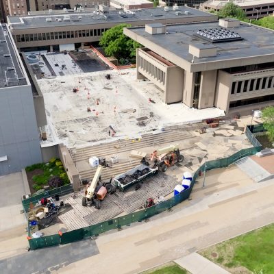 Copilot said: Aerial view of a university building and plaza under construction, with excavation work, construction equipment, fencing, and surrounding campus buildings.