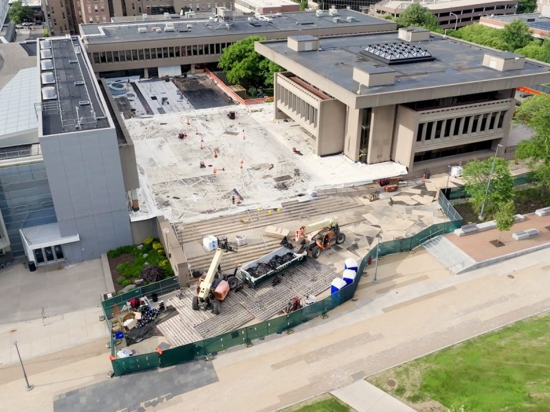 Copilot said: Aerial view of a university building and plaza under construction, with excavation work, construction equipment, fencing, and surrounding campus buildings.