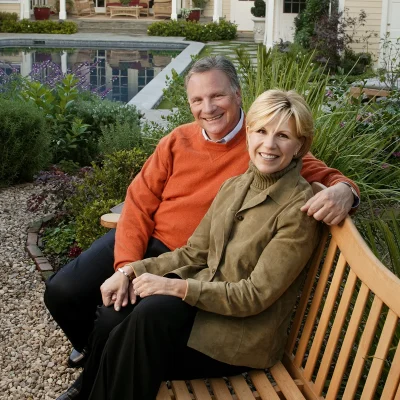 A couple sit together on a wooden bench in a landscaped garden with a pool and light-yellow house in the background.