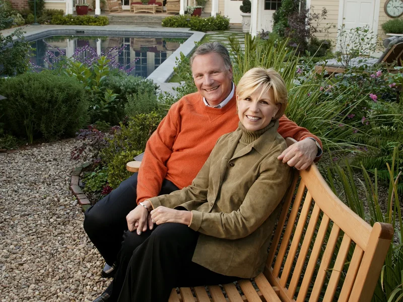 A couple sit together on a wooden bench in a landscaped garden with a pool and light-yellow house in the background.