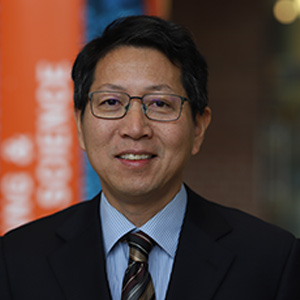 Portrait of Quinn Qiao, a man wearing glasses, a dark suit, and a striped tie, standing indoors with an orange banner reading 'SCIENCE' in the background.