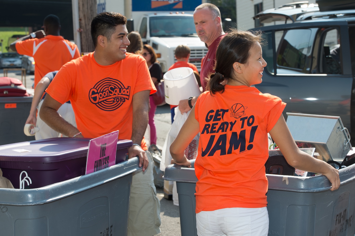 Volunteers in bright orange shirts move storage bins and belongings during a campus move-in event.