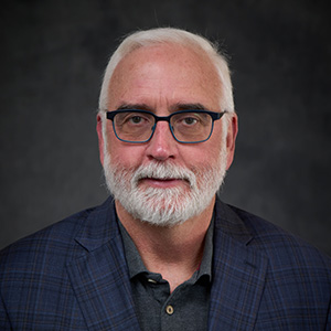 Professional headshot of a man with white hair, a white beard, and glasses, wearing a navy plaid blazer over a dark polo shirt, photographed against a dark gray background.
