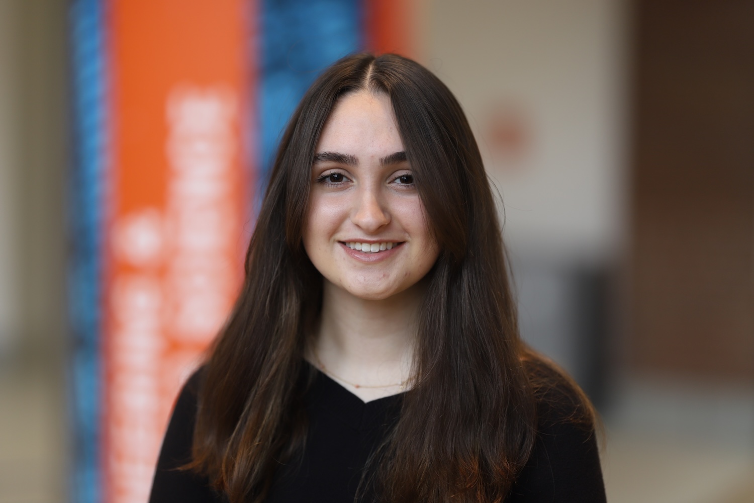 Professional indoor headshot of a person with long dark hair wearing a black top, with a softly blurred background.