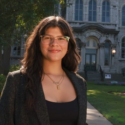 Young woman with long dark hair and glasses smiling outdoors in front of the Hall of Languages, wearing a black top and blazer.