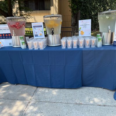 Table with three infused-water dispensers (strawberry, citrus, cucumber), stacked cups, and wellness/hydration signs at an outdoor campus event.