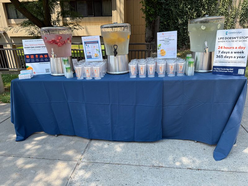 Table with three infused-water dispensers (strawberry, citrus, cucumber), stacked cups, and wellness/hydration signs at an outdoor campus event.