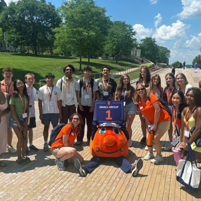 A group of orientation students pose outdoors with Otto the Orange mascot beside a "Small Group 1" sign.