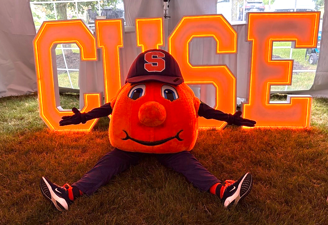 Otto the Orange mascot sits on grass in front of glowing “CUSE” letters, arms spread wide.