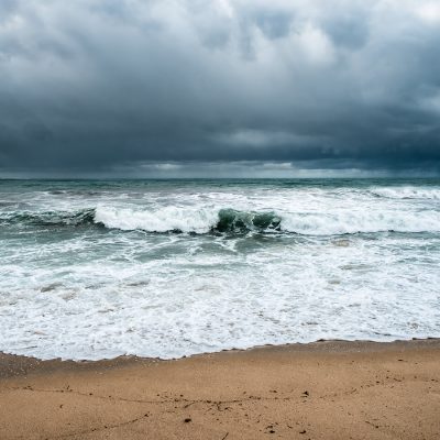 Dark storm clouds hang over a turbulent teal ocean with white-capped waves breaking onto a sandy beach.