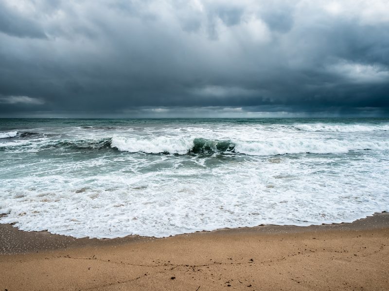 Dark storm clouds hang over a turbulent teal ocean with white-capped waves breaking onto a sandy beach.
