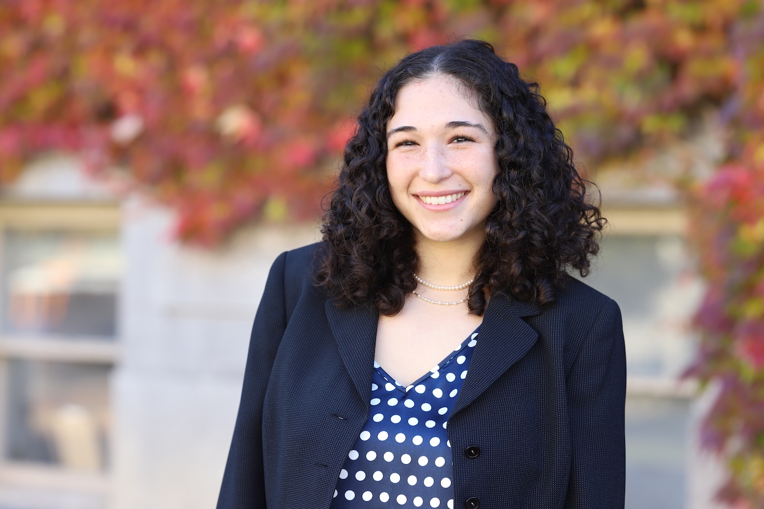 Professional outdoor headshot of a person in a dark blazer and polka-dot blouse, standing in front of colorful autumn foliage.