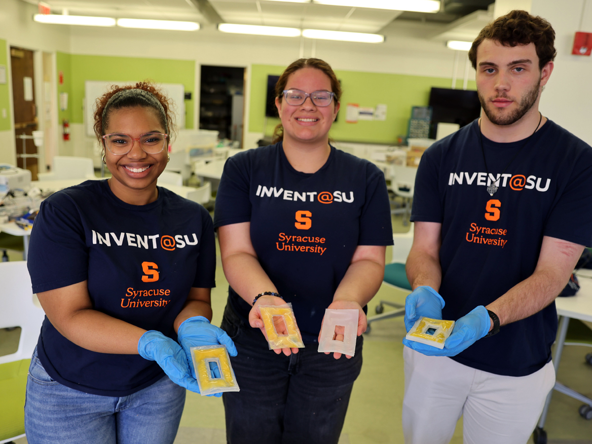 Three Invent@SU students in matching shirts hold prototype devices in a lab.
