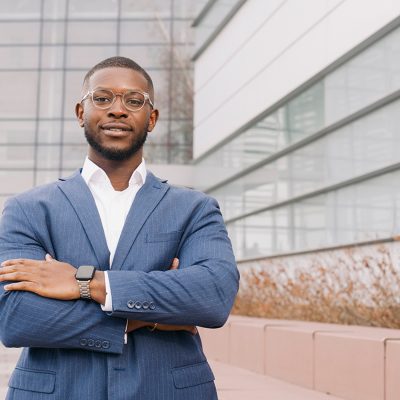 A man with glasses and a beard, wearing a navy pinstripe suit with a white open-collar shirt, stands with arms crossed in front of a modern glass office building, with a smart watch visible on his wrist.