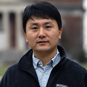 Headshot of a man with short black hair, wearing a light blue collared shirt under a navy fleece jacket, standing outdoors with a blurred building and trees in the background.