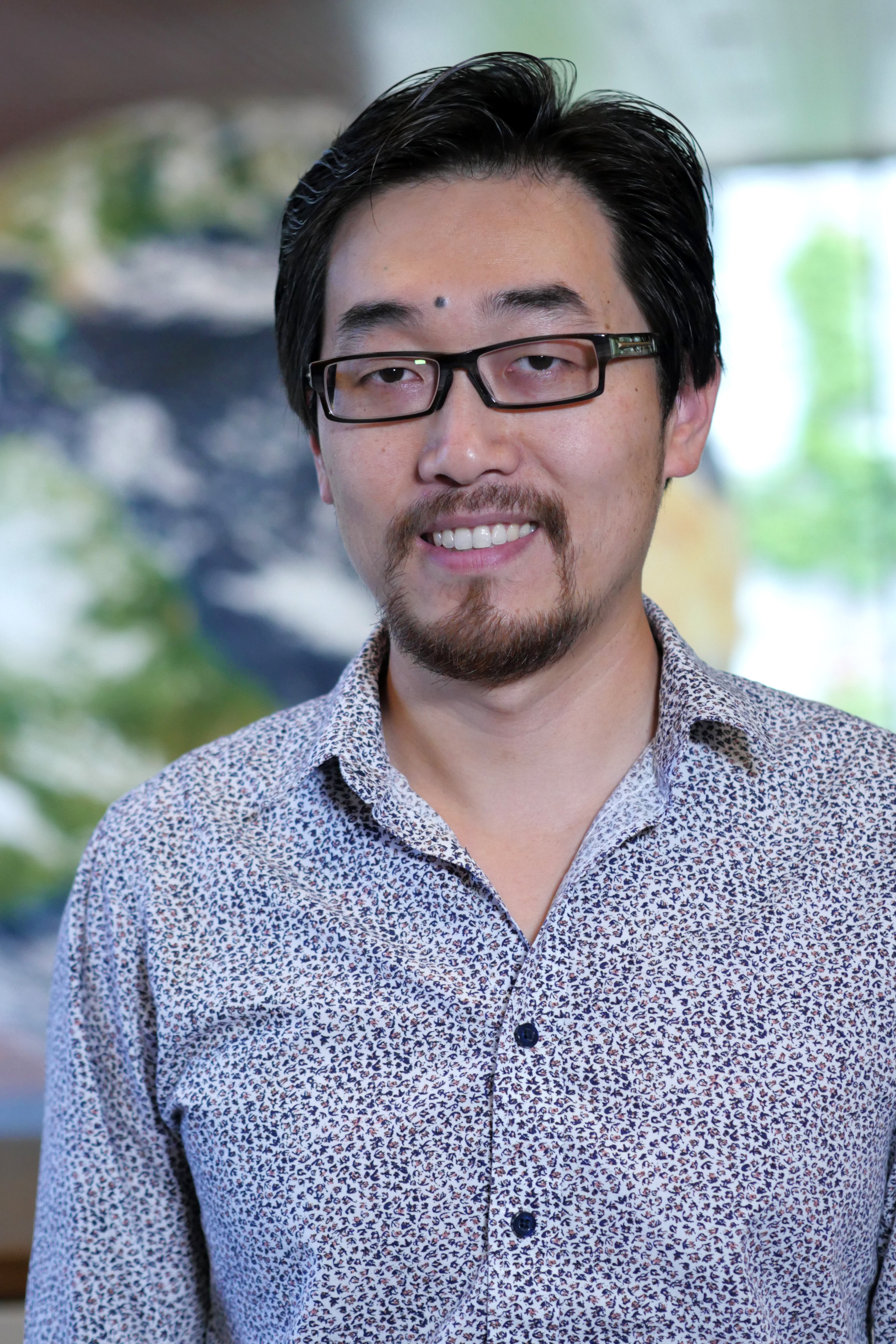 Head-and-shoulders portrait of a person wearing glasses and a patterned button-down shirt, standing in front of a blurred map-like background.