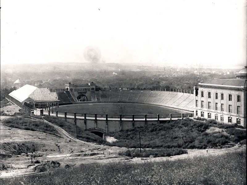 Historic photograph of a large outdoor stadium beside a university building, viewed from a hillside overlooking the surrounding city.