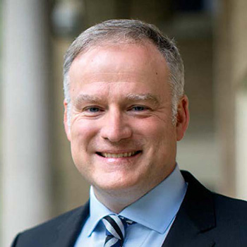 Head-and-shoulders portrait of a person in a suit and tie, facing the camera, with blurred columns in the background.