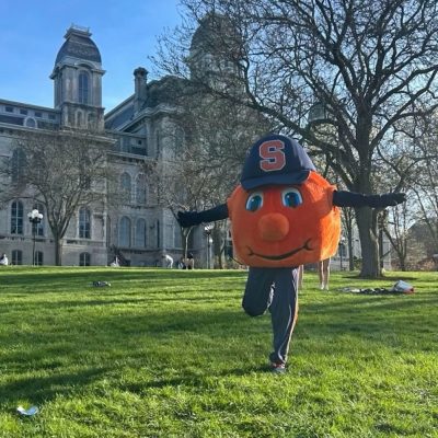 Otto the Orange mascot balances on one leg on a campus lawn in front of a historic stone building.