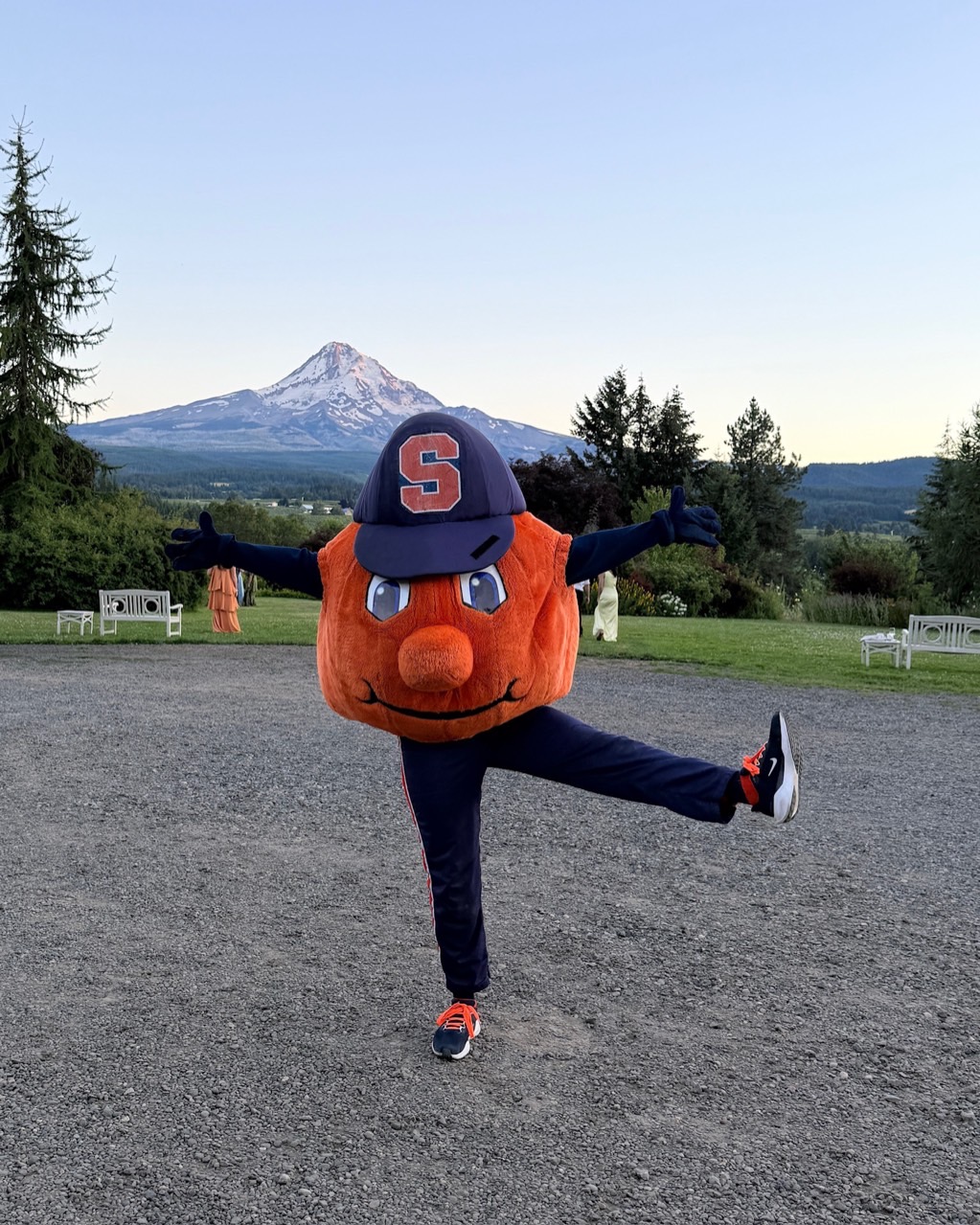 Otto the Orange mascot poses with arms outstretched and one leg raised, with a mountain landscape in the background.