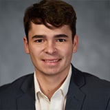 Professional headshot of Dylan LeHouillier, a man with short brown hair, smiling, wearing a navy blazer with a light blue shirt against a gray background.