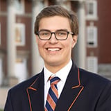 Professional headshot of Evan Greenberg, a man with short blond hair and glasses, smiling, wearing a navy blazer with a striped tie, with a brick campus building in the background.