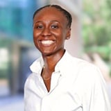 Professional headshot of Kelsey Davis, a woman with short black hair, smiling, wearing a white collared top against a blurred outdoor background.