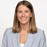 Professional headshot of Makenzie Mertikas, a woman with straight blonde hair, smiling, wearing a light gray blazer against a white background.