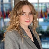 Photo of Megan Edenfeld, a woman with wavy strawberry-blonde hair, smiling, wearing a gray blazer over a black top, with international flags blurred in the background.