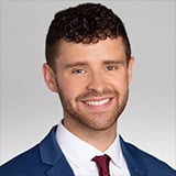 Professional headshot of Sam Cestari, a man with curly brown hair and a beard, smiling, wearing a navy suit with a white shirt and burgundy tie against a gray background.