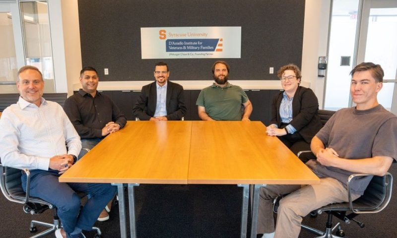 Six people sit around a wooden conference table, with a sign reading 'Syracuse University D'Aniello Institute for Veterans & Military Families' mounted on the wall behind them.