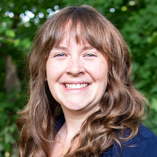 Headshot of a person with long wavy hair outdoors, with green foliage in the background.