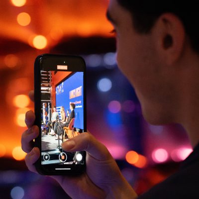 Person films a "Launch Night" panel discussion on their phone, colorful lights blurred behind.