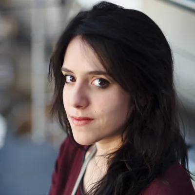 Head-and-shoulders portrait of a person with dark hair, looking toward the camera, against a softly blurred outdoor background.