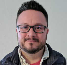 Headshot of a man with short dark hair and glasses wearing a dark jacket over a light patterned collared shirt against a plain light background